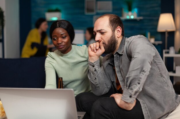 Multi-ethnic friends sitting on couch watching online video on laptop enjoying spending time together during wekeend party. Group of people hanging out, relaxing in living room. Friendship concept