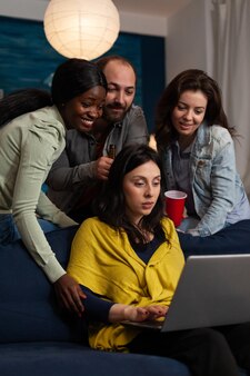 Multi-ethnic friends smiling while watching online comedy series on laptop computer during entertainment movie home night. group of people enjoying hanging out together. friendship concept