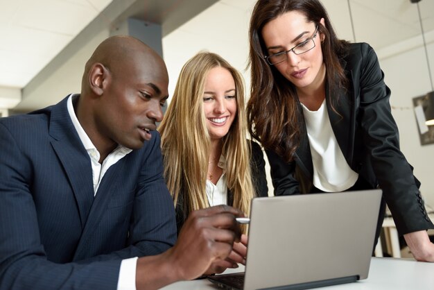 Многоэтническая группа из трех бизнесменов, встречающихся в современном офисе. Две женщины Кавказа и черный человек в костюме, глядя на портативный компьютер.