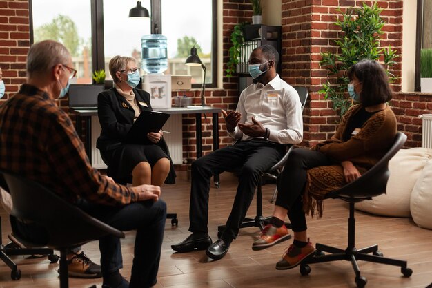 Multi ethnic people discussing about addiction at support group therapy, during covid 19 pandemic. Patients with face masks at aa meeting to cure mental health problems with specialist.