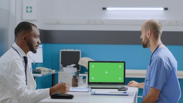 Многоэтническая команда обсуждает медикаментозное лечение на макете ноутбука с цветным ключом с зеленым экраном и изолированным дисплеем. Афро-американский врач-терапевт объясняет медицинскую экспертизу