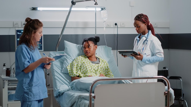 Multi ethnic team of specialists consulting young patient with disease in hospital ward. Medical assistant and doctor giving support and helping sick woman with recovery. Healthcare workers