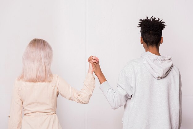 Multi ethnic teenage couple standing against white wall holding each other's hand