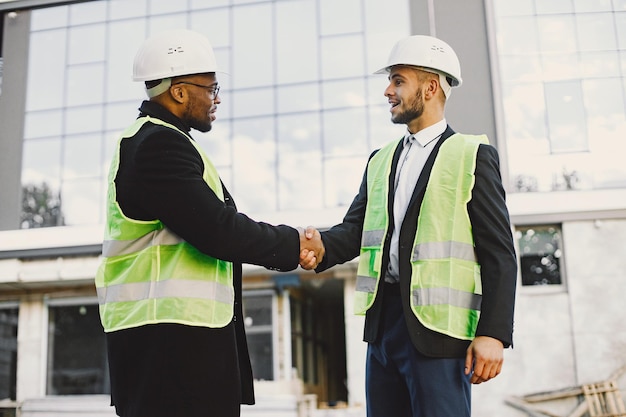 Multi racial builders handshaking outdoors. Wearing uniform, talking about new glass building. Working on the poject. City infrastructure