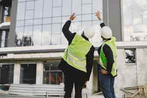 Foto gratuita costruttori multirazziali in piedi all'aperto, vista posteriore. indossando l'uniforme, parlando di un nuovo edificio di vetro. lavorare sul progetto. infrastrutture cittadine