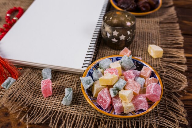 Multicolor cubes of rakhat-lukum in plate with blank white spiral notebook and candle holder on sack tablecloth