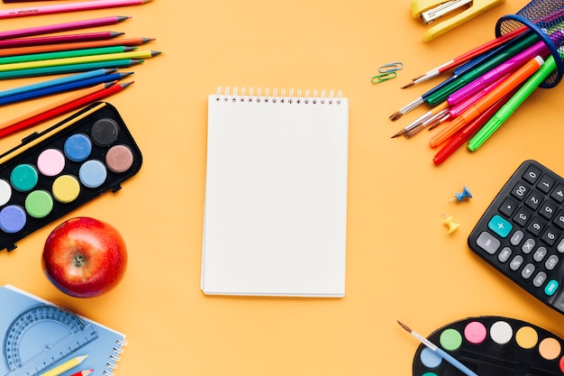 Multicolor school stationery scattered around blank notepad on yellow desk