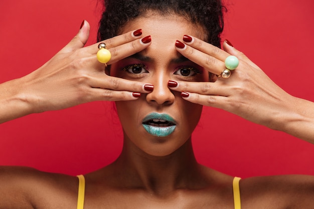 Free photo multicolor young mulatto woman with trendy makeup looking on camera through fingers with rings, over red wall