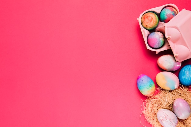 Multicolored easter eggs in nest and carton box on pink backdrop