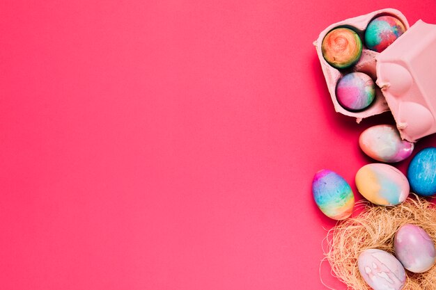 Multicolored easter eggs in nest and carton box on pink backdrop