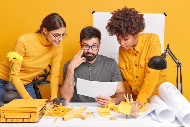 Free photo multicultural partners cooperate on design project discuss ideas look attentively at illustration on paper brainstroming together at blueprint sketches pose in coworking space. diverse professionals