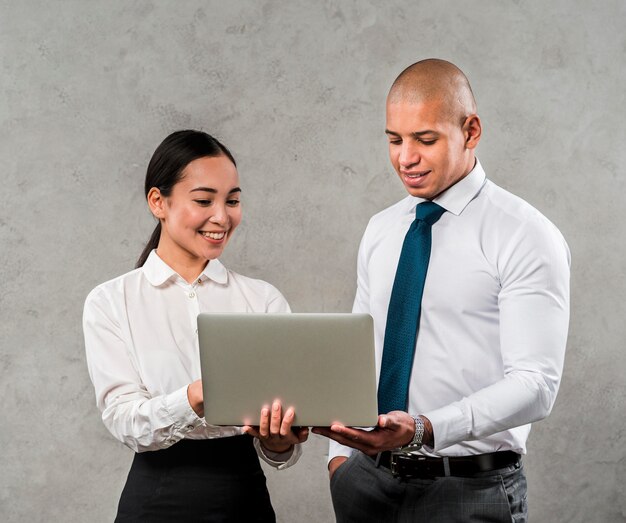 Multiethnic businessman and businesswoman looking at laptop standing against grey wall