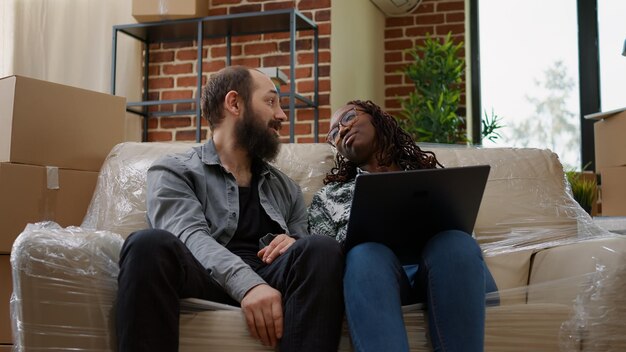 Multiethnic couple finding home decor inspiration on laptop, doing online shopping to move in new rented apartment flat. Unpacking house furniture and belongings for new beginnings.