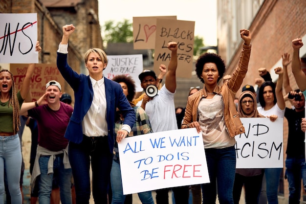 Multiethnic crowd of people with raised fists protesting for human rights while carrying antiracism placards through city streets