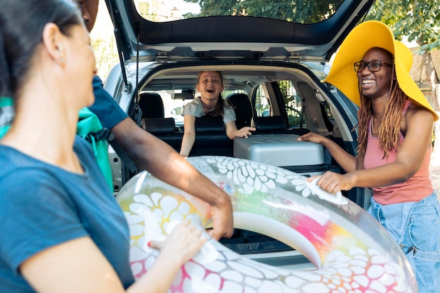 Free photo multiethnic family and friends loading bags in car, preparing baggage in automobile. travelling to seaside with diverse people, putting luggage and inflatable in vehicle, leaving on vacation.