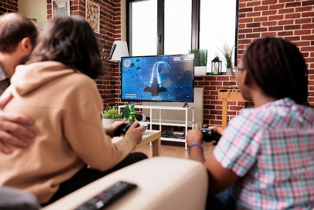 Multiethnic friends sitting at home in living room while playing space videogame simulator together. Adult people enjoying fun leisure activity on modern gaming console device while having beer and sn