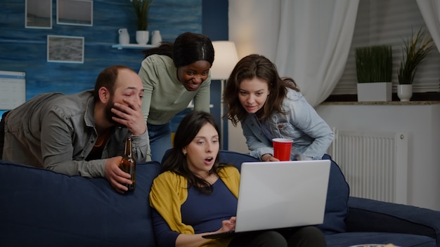 Многонациональные друзья смотрят интересный комедийный фильм на портативном компьютере, расслабляясь на диване, пьют ...