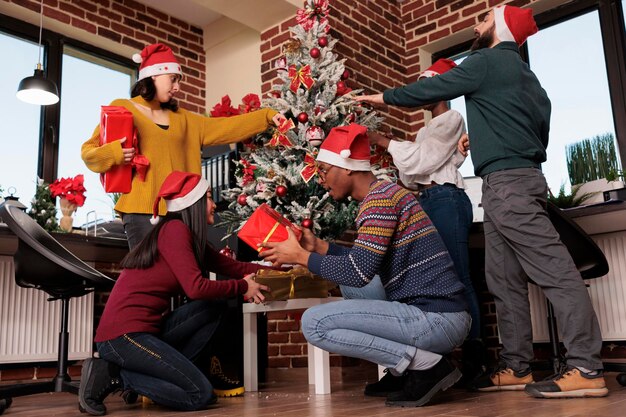 Многоэтническая группа коллег украшает офис праздничными украшениями и елкой, дарит подарки и подарки. Празднование рождественской праздничной традиции зимой на рабочем месте.