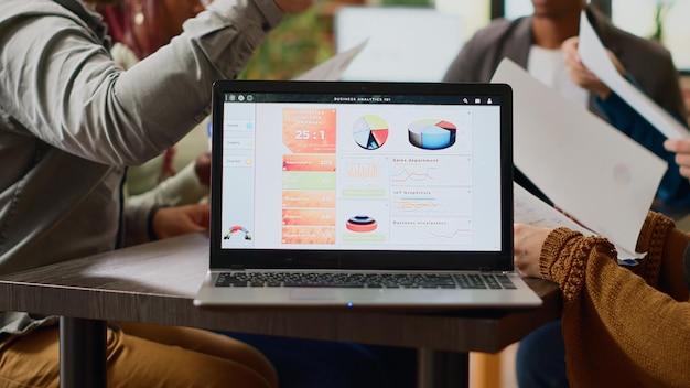 Multiethnic group of coworkers analyzing business charts and infographics data on laptop software. People doing teamwork collaboration to create paper reviews and professional report. Close up.
