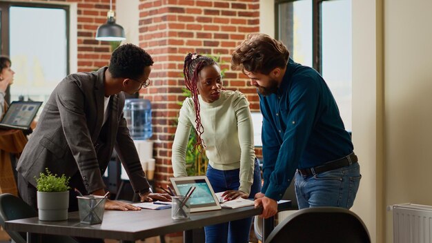 Multiethnic group of coworkers analyzing information on charts, using paperwork and online research on digital tablet. People planning business report with data analysis and ideas.