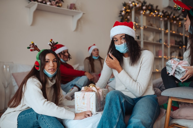 Gruppo multietnico di amici in cappelli di babbo natale sorridenti e in posa per la telecamera con doni nelle mani. il concetto di celebrare il nuovo anno e il natale sotto le restrizioni del coronavirus. vacanza in quarantena