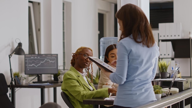 Multiethnic group of people analyzing annual report on files and computer, checking online data in business meeting. Young startup team discussing about report strategy in coworking space.