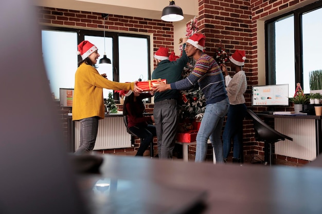 Free photo multiethnic group of people exchanging holiday gifts at christmas celebration in business office. colleagues giving presents in workplace decorated with festive xmas ornaments and lights.