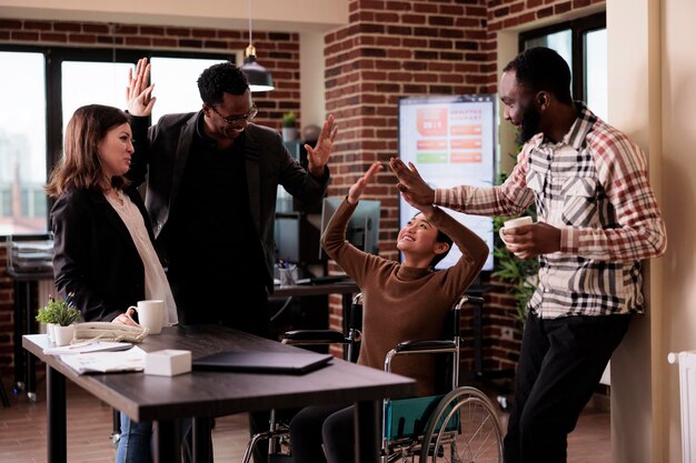 Multiethnic group of people having fun at work on break, laughing and enjoying conversation with paralyzed employee. Woman living with physical chronic disability in wheelchair at job.