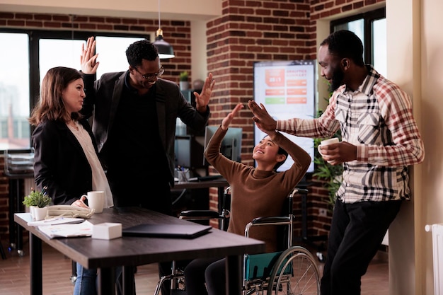 Multiethnic group of people having fun at work on break, laughing and enjoying conversation with paralyzed employee. Woman living with physical chronic disability in wheelchair at job.