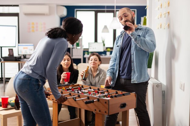 Multiethnic group of people playing at foosball table game, meeting for drinks in office after work hours. Man and women drinking beer from bottles and eating snacks at celebration.