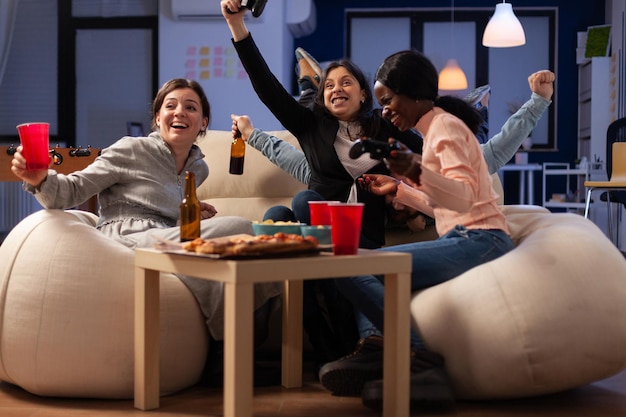 Multiethnic group of people winning video games play with joystick on console, meeting in office for drinks after work hours. Man and women celebrating game win and drinking beer.