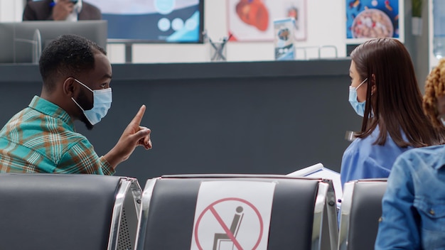 Multiethnic group of people with face masks sitting in waiting area at hospital reception lobby. Nurse and african american man talking about healthcare service during covid 19 epidemic.