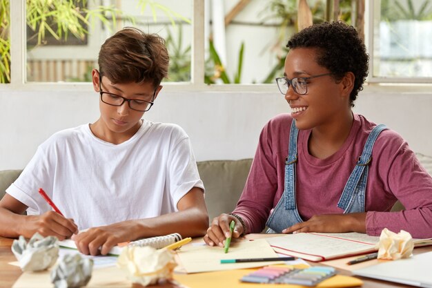 Multiethnic groupmates write common essay, sit at table with papers and notepad, dressed in casual clothing. Asian boy recieves consultancy from dark skinned friend, check information from documents