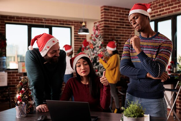 Multiethnic team of coworkers using laptop on christmas, working on project in office decorated with festive ornaments and xmas tree. People with santa hat doing teamwork during holiday.