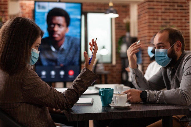 Foto gratuita team multietnico con maschera medica protettiva per prevenire l'infezione da covid19 che discute della strategia di marketing con il manager remoto durante la videochiamata online nell'ufficio di avvio. chiamata d'affari