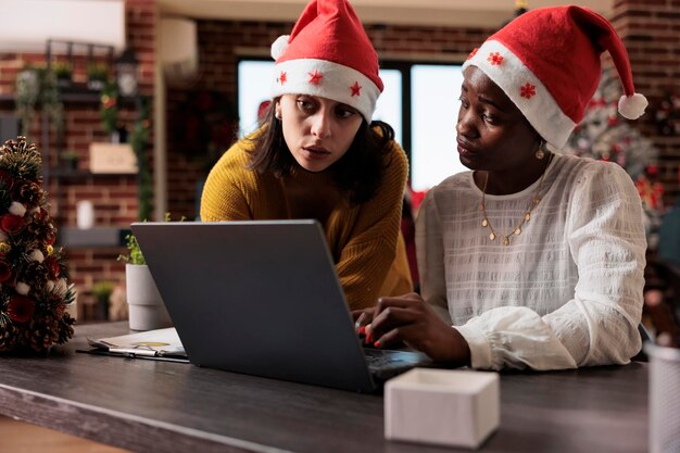 산타 모자를 쓴 다민족 여성 팀은 크리스마스 장식과 장신구로 가득 찬 축제 사무실에 앉아 노트북에서 사업을 하고 있습니다. 팀워크를 하고 휴가를 축하합니다.