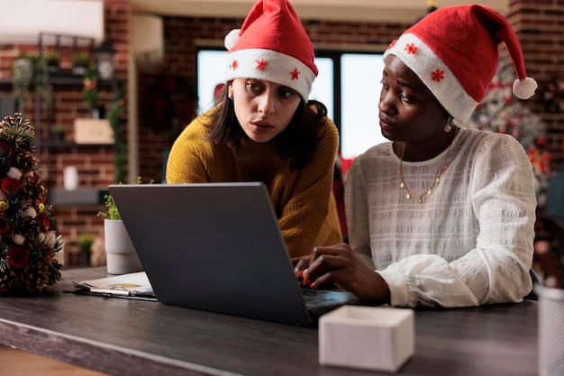 Многонациональная команда женщин в новогодней шапке, работающих по делам за ноутбуком, сидящих в праздничном офисе, наполненном рождественскими украшениями и украшениями. Работа в команде и празднование праздника.