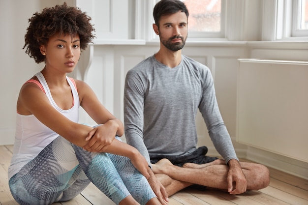 Multiethnic woman and man meditate together on floor, have good flexibility, practice yoga in domestic setting, look with confident expression, feel relaxed. People and concentration