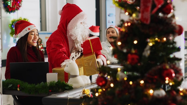 Multiethnic workers receiving gifts from colleague pretending to be Santa in xmas decorated office Employee wearing costume surprising company personnel with presents during winter holiday season