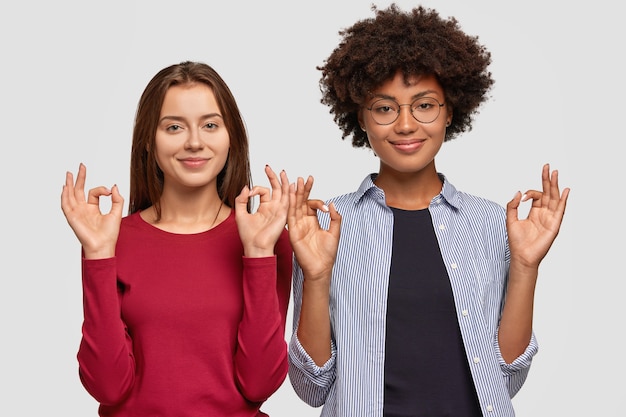 Multiethnic young women recommend you something, gesture indoor and show okay sign with both hands