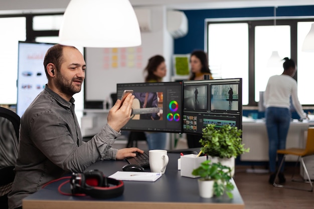 Multimedia creator artist talking on videocall with client holding smartphone editing video footage sitting in creativity production agency. videographer using post production software