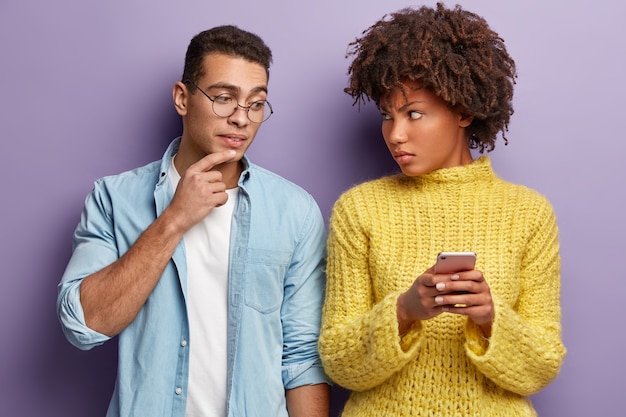 Free photo multiracial couple get news on cellular. angry woman asks boyfriend not to peek in cell phone, looks in dissatisfaction, wears yellow jumper. curious caucasian man in spectacles has interest