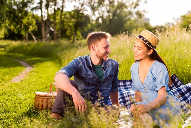 Multiracial enamored couple sitting on checkered plaid on grassy meadow