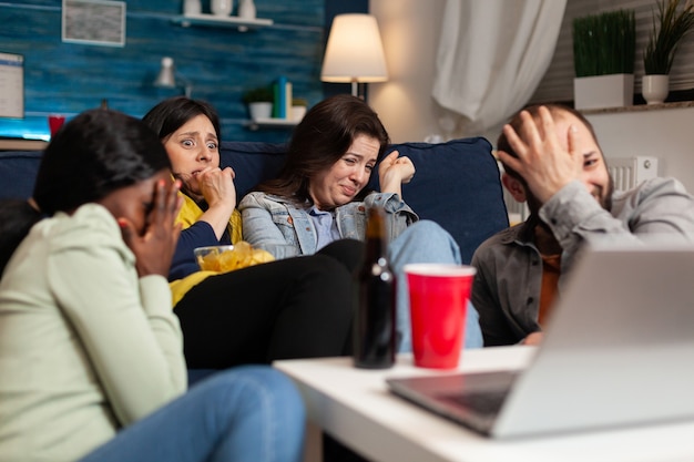 Multiracial friends chilling on sofa sitting in front of television watching horror thriller movie during night series