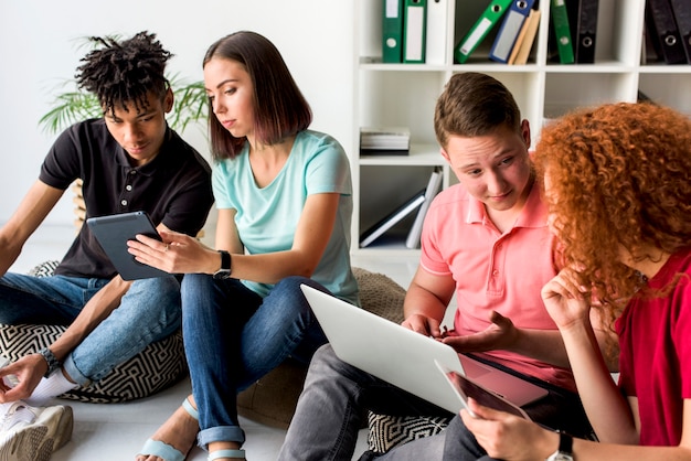 Multiracial friends using electronic gadgets sitting on floor having discussion