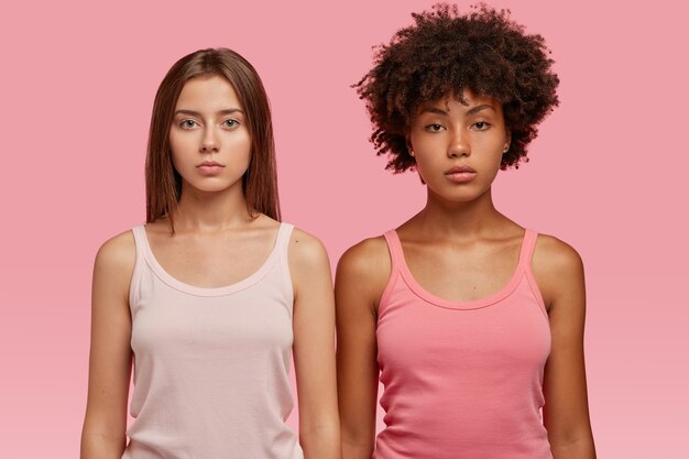캐주얼 조끼를 입은 다민족 여성들은 분홍색 벽에 진지하게 서서 함께 스포츠 훈련을 할 것입니다.