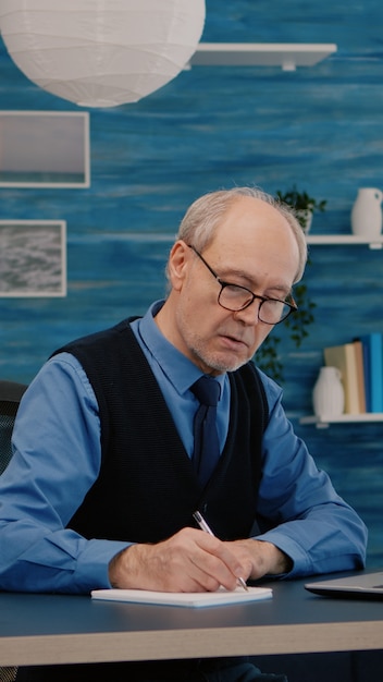 Multitasking retired man reading on laptop and writing on notebook working from home