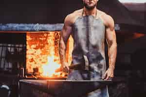 Free photo muscular blacksmith in protective apron at his workshop with hammer in his hands. there are fire at background.