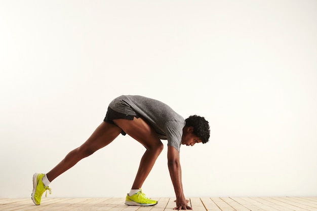 Muscular dark skinned sprinter in gray shirt, black shorts and neon yellow sneakers getting into starting position shot from the side against white wall