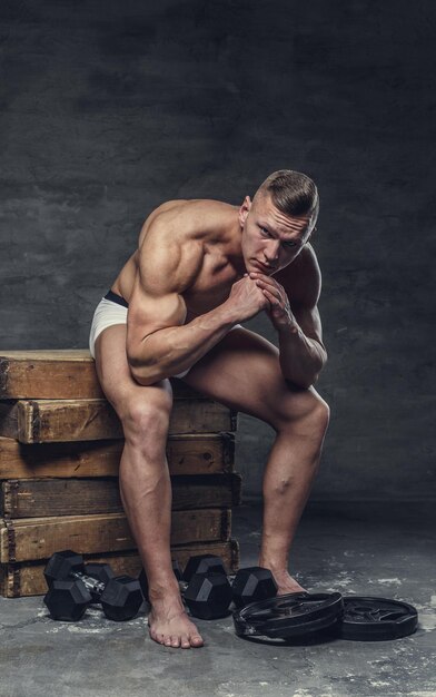 Muscular guy in a white panties sits on a wooden box.